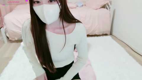 Media: Video of a young Asian woman with long brown hair, wearing a white mask, long-sleeve top, and black pants, kneeling on a white rug in a pastel-pink bedroom.