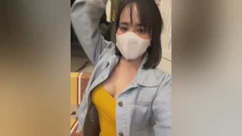 Media: Video of an Asian woman with medium skin tone, straight black hair, and a white face mask, wearing a yellow dress, light blue denim jacket, and a brown purse. She is indoors, possibly in a public restroom, with blurred background.