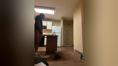 Media: A video shows a woman with a dark complexion and short hair in a dimly lit, cluttered hotel room. She is barefoot, wearing a black top and no pants, standing by a wooden dresser. A white refrigerator and microwave are visible in the background.