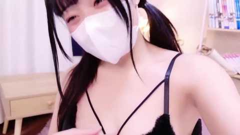 Media: Video of an East Asian woman with long black hair, wearing a white mask, black lace bra, and large gold earrings. She's indoors, with a white curtain and bookshelf in the background.