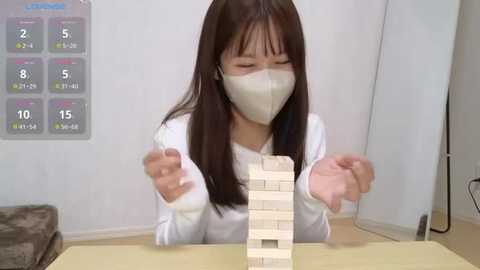 Media: Video of an East Asian woman with long brown hair and a white mask, building a wooden Jenga tower. Background shows a calendar and white wall.