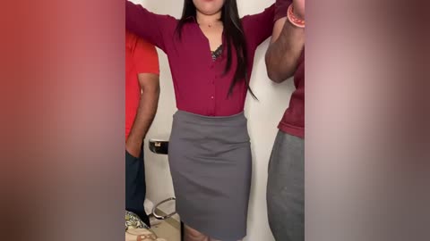 Media: Video of a woman with long black hair, wearing a maroon cardigan over a grey pencil skirt, posing in a room with a white wall, a red shirt on the left, and a man in a maroon shirt on the right.