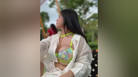 Media: A video of a woman with long black hair, wearing a white shirt over a neon green bra with floral patterns, sitting outdoors, blurred background.