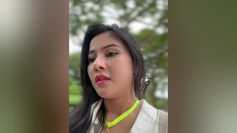 Media: Video of a young woman with long black hair, wearing a neon green choker, standing outdoors against blurred greenery.