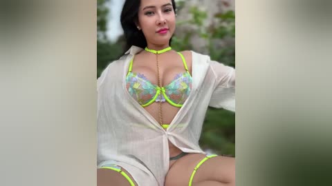 Media: Video of a Latina woman with long dark hair, wearing neon yellow lingerie and a sheer white blouse, seated outdoors with blurred green foliage in the background.