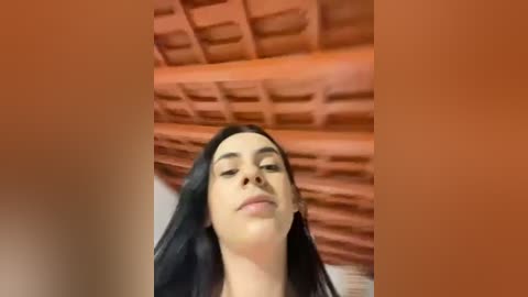 Media: Video of a young woman with long black hair, wearing a white top, standing under an orange-tiled roof with a wooden beam structure. She has a calm expression.