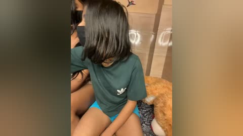 Media: Video of a young woman with long black hair, wearing a green Adidas T-shirt and blue shorts, sitting on a plush toy, in a cozy, warmly lit room with a washing machine and dryer in the background.