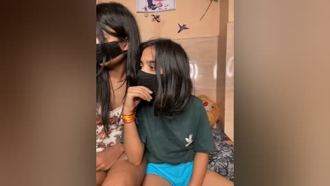 Media: Video of a young woman with long black hair, wearing a floral dress, and a mask, sitting on a bed next to a young girl with short black hair in a green shirt and blue shorts.