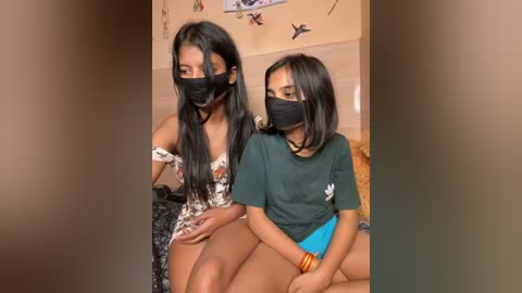 Media: Video of two young women with long black hair, wearing face masks, in casual clothes, seated on a bed. Background shows a beige wall with butterfly stickers.