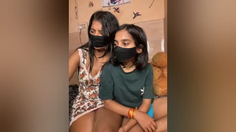 Media: Video of two young women with medium brown skin, wearing masks, black tops, and floral dresses. They sit on a bed in a room with beige walls and bird stickers.