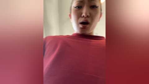 Media: Video of an Asian woman with a light skin tone and short black hair, wearing a red dress, standing in front of a blurred background, mouth slightly open, looking surprised or shocked.