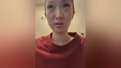 Media: A video of a young East Asian woman with light skin, wearing a red shirt, with long, dark hair pulled back, and expressive green eyes, standing indoors with blurred beige walls.