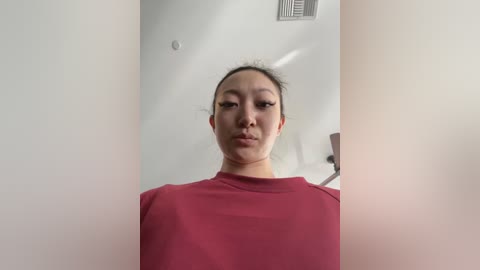 Media: A video of an Asian woman with light skin and straight black hair tied back, wearing a red t-shirt. The background is a white wall with a small vent and a blurred lamp.