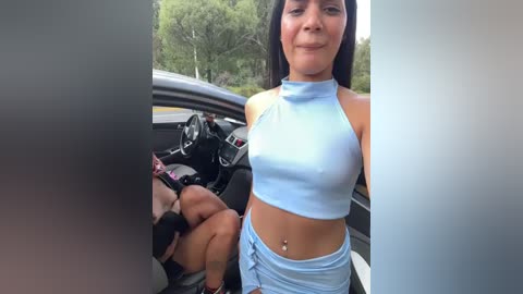 Media: Video of a Latina woman with medium skin tone, wearing a light blue halter top and matching high-waisted shorts, seated in a car with a muscular man's arm around her.