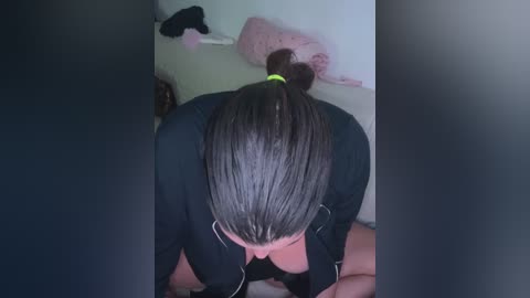 Media: A video shows a woman with straight, dark hair tied back in a ponytail, wearing a black shirt, kneeling on a bed with a pink stuffed animal and clothes hanging on a wall in the background.