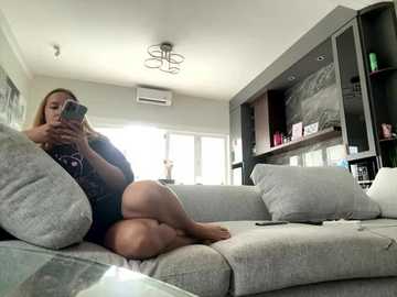 Media: Video of a woman with light skin and long blonde hair, wearing a dark t-shirt and shorts, sitting on a grey couch in a modern living room with large windows, shelves, and an air conditioner.