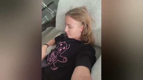 Media: Video of a blonde woman in a black t-shirt with pink cartoon character, reclining on a beige couch, eyes closed, holding a phone, indoor setting.