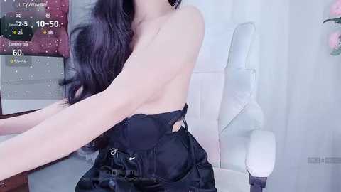 Media: Video of a topless Asian woman with long black hair, wearing black pants, sitting on a white chair in a minimalist room.