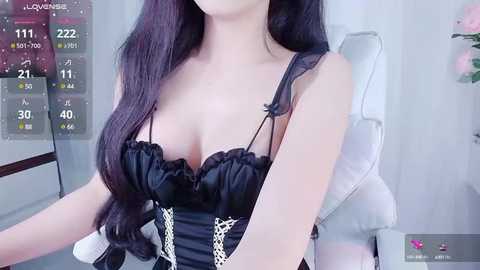 Media: Video of an Asian woman with long, wavy black hair, wearing a revealing black corset with white lace, sitting on a white leather chair. Background shows digital stats overlay and a pink flower vase.