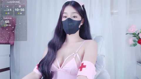 Media: Video of an East Asian woman with long black hair, wearing a pink nurse outfit with red cross, black face mask, and a white chair in a room with pink flowers.