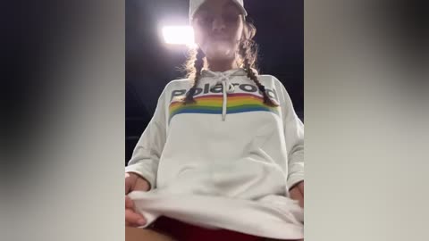 Media: A video of a young woman with long, braided hair wearing a white \"Polo\" hoodie and red shorts, standing in a dimly lit room with a bright light source behind her.