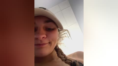 Media: Video of a young woman with light skin, curly brown hair, wearing a beige hat, smiling slightly, with a spider crawling on her face. Background is a white wall and ceiling.