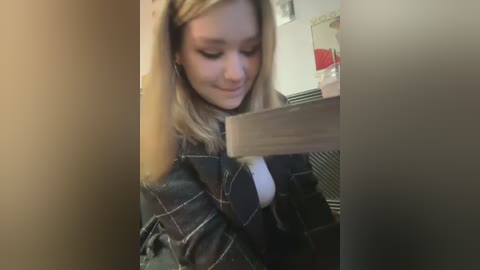 Media: A video shows a young blonde girl with a plaid shirt, smiling while holding a wooden stick, in a dimly lit room with a fridge and red items in the background.