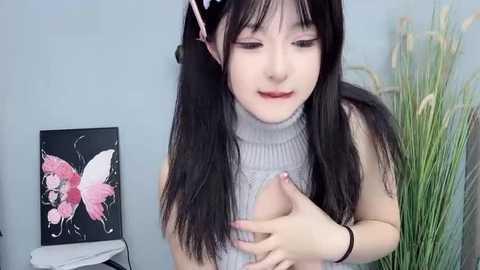 Media: A video of a young East Asian woman with long black hair, wearing a grey turtleneck sweater, covering her breasts. She has fair skin and is in a room with a floral painting and greenery.