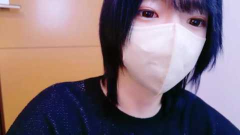 Media: Video of a person with short, straight black hair, wearing a white surgical mask, black sweater, and standing in front of a wooden door.