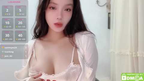 Media: Video of an Asian woman with long black hair, fair skin, wearing a white lace bra, slightly exposing her cleavage, in a modern, well-lit room.