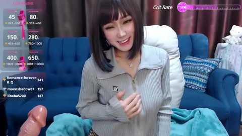 Media: Video of a smiling Asian woman with straight black hair, wearing a grey sweater, holding a large erect penis in her left hand.