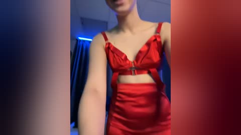 Media: Video of a fair-skinned woman in a red satin lingerie set with a plunging neckline and high-waisted skirt, standing indoors against a blue-lit background.