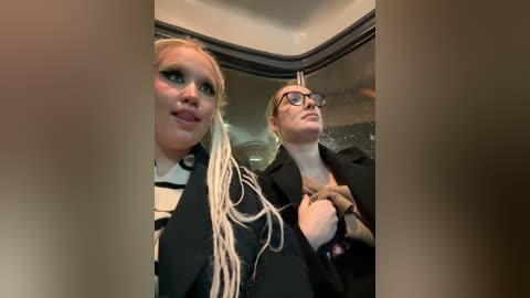 Media: Video of two women with long blonde hair and glasses, one in a black jacket, the other in a black top, standing inside a car.