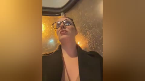 Media: Video of a woman with glasses, wearing a black jacket over a low-cut top, looking up through a rain-streaked window. Background shows blurred street lights.