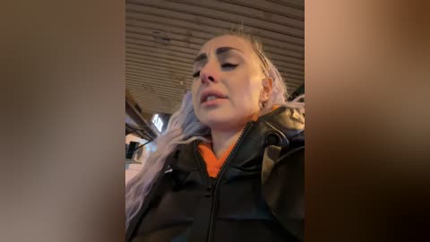 Media: Video of a blonde woman with closed eyes, wearing a black leather jacket, inside a vehicle with a wooden ceiling and blurred background.