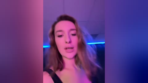 Media: Video of a woman with light skin and long, wavy, blonde hair, wearing a black top, in a dimly lit room with purple and blue neon lighting.