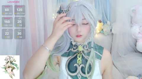 Media: Video of a pale-skinned woman in a white dress with intricate green patterns, adorned with a silver bracelet and a floral headpiece. She has long, silver hair and a serene expression, with a background featuring a plush toy and sheer curtains.