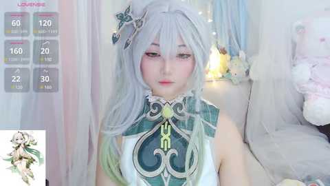 Media: Video of a young Asian woman in a white and green cosplay dress with a flower headpiece, standing in a soft-lit bedroom with pastel decor.