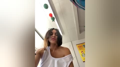 Media: A video of a woman with shoulder-length dark hair, wearing a white off-shoulder top, standing in a brightly lit room with colorful decorations and posters on the wall.