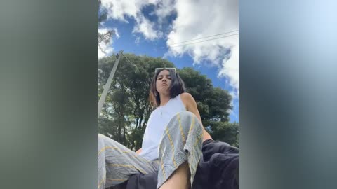 Media: Video of a young woman with medium skin tone and shoulder-length dark hair, wearing a white tank top and gray pajama pants, sitting on a dark couch outside under a partly cloudy sky with trees in the background.