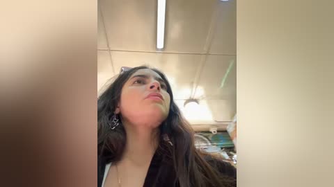 Media: Video of a young woman with long, dark hair and fair skin, wearing a black jacket, looking upward with a thoughtful expression in a dimly lit room with fluorescent lights and cluttered shelves in the background.