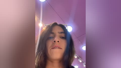 Media: A video of a young woman with long brown hair, light skin, and a slight smile, taken from a low angle, showing her face prominently. The background features a pink ceiling with string lights and a blurred, warm color palette.