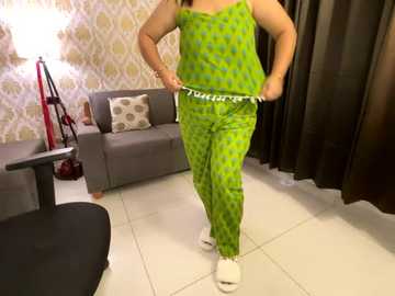 Media: Video of a person with medium skin tone, wearing a green, polka-dotted pajama set, standing in a modern living room with a grey sofa, patterned wallpaper, and beige tiles.