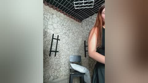 Media: Video of a woman in a black dress standing in a small, dimly lit room with cracked white walls, a grey stool, and a metal ladder.