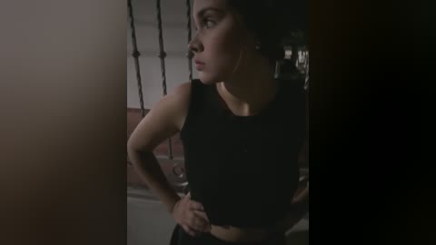 Media: Video of a woman with medium skin tone, dark hair, wearing a black sleeveless top, looking contemplative, standing indoors with a dimly lit background.