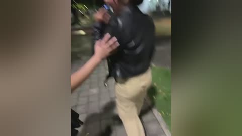 Media: A blurry video shows a person in a black leather jacket and beige pants being restrained by two hands, standing on a stone-paved path with green grass and foliage in the background.