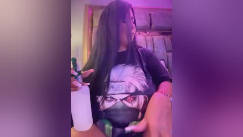 Media: Video of a person with long, dark hair, wearing a black t-shirt with a Naruto graphic, holding a white plastic bottle. The background includes a cushioned chair and a window with curtains.