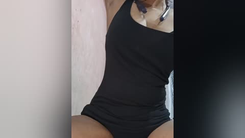 Media: A video of a woman in a black tank top and matching panties, taken indoors against a white and black backdrop, showing her torso and upper thighs.