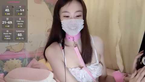 Media: A video of an Asian woman with long black hair, wearing a white lace bra, pink mask, and pink collar, holding a phone, in a pastel bedroom.