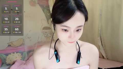 Media: A video of a young East Asian woman with fair skin, dark hair in a bun, wearing headphones with a pink strap, sitting in a room with a pink bed and cartoonish wall art.